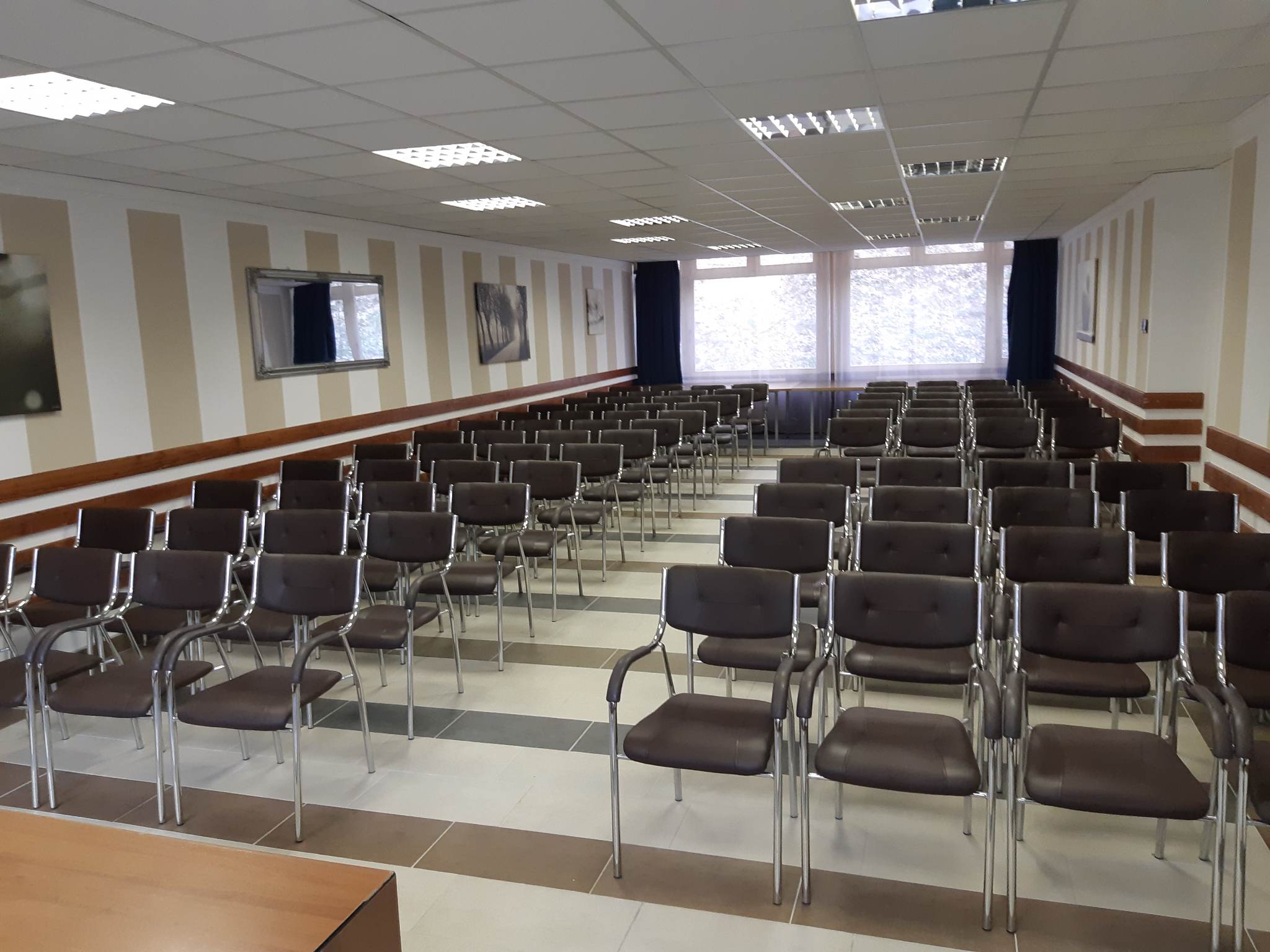 Rendezvényterem sorokba rendezett, fémvázas székekkel, a falra szerelt képernyővel.